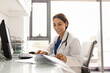 © fizkes - Young 35s Middle-Eastern female doctor in uniform working seated at workplace table with papers and computer, reviewing medical documents, check patient records or analyzing clinical reports on paper