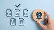 © shahabudin - A human hand holds a wooden quality assurance badge with a checkmark next to document icons featuring completed checklists on a light blue background