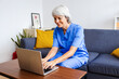 © Xavier Lorenzo - Portrait of senior healthcare worker in blue scrubs working on laptop sitting on sofa at home. Technology and healthcare business concept.