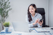 © MINAE - Asian businesswoman working in office, smiling and using a smartphone while looking at financial reports and a laptop, analyzing company data and business strategies