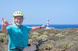 © luciano - Senior man enjoying a cycling break on a coastal trail with a red and white lighthouse and the Atlantic Ocean in the background in Tenerife, Canary Islands. Active lifestyle and healthy retirement