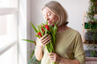 © Tatyana Gladskih - Happy Mother's Day and Women's Day, Charming woman with a bouquet of tulips in the living room by the window, woman enjoys smelling flowers, smell of spring and freshness.