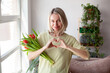 © Tatyana Gladskih - Happy woman enjoy bouquet of tulips, she shows a heart sign in gratitude, Housewife enjoying a bunch of flowers and interior of living room