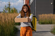© wifesun - Woman courier delivering a cardboard parcel to a smiling customer outside a home, wearing striped shirt and orange pants, holding a yellow insulated delivery bag, urban logistics worker
