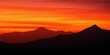 © antishock - Dramatic sunset over mountains. Vivid orange sky contrasts dark silhouettes. City lights glimmer below peaks. Peaceful, serene view.