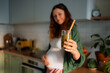 © wifesun - Pregnant woman offering a glass of green smoothie with a wooden straw, portraying healthy eating habits and conscious wellness choices during pregnancy in her home kitchen
