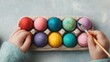 © Intelligent Horizons - Close up of a young child s delicate hands using a paintbrush to decorate vibrant multi colored Easter eggs arranged in a cardboard holder against a simple neutral background