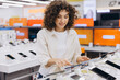 © anatoliycherkas - Woman exploring digital devices in electronics store, shopping for new tablet or smartphone, consumer technology