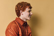 © SHOTPRIME STUDIO - Redhead man with curly hair wearing a brown jacket smiling and looking sideways on beige background in studio portrait with casual style and warm colors.