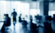© birulaut - A blurred office scene featuring individuals working at desks in an ambient environment, with sunlight streaming through large windows.