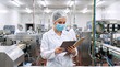 © Vasiliy - Female quality control inspector in a white lab coat and mask. Industrial worker holding a clipboard in a pharmaceutical factory. Manufacturing plant production line
