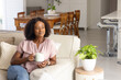 © Wavebreak Media - African woman sitting on light beige sofa holding white mug in living room with throw pillows