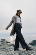 © SHOTPRIME STUDIO - Young woman wearing stylish coat and cap walks confidently on rocky beach near calm sea under cloudy sky. Concept of fashion, autumn, and outdoor lifestyle with natural light.
