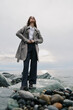 © SHOTPRIME STUDIO - Confident woman wearing long coat and wide pants stands on rocky shore near ocean under cloudy sky. Concept of fashion, autumn style, and outdoor lifestyle with natural coastal environment.