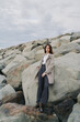 © SHOTPRIME STUDIO - Young woman in grey coat and dark pants stands on rocky shore under cloudy sky. Casual fashion and outdoor lifestyle concept with natural landscape and relaxed mood by the sea.
