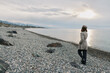 © SHOTPRIME STUDIO - Woman in coat and hat walks alone on rocky beach during cloudy sunset. Calm sea and distant mountains create peaceful and contemplative mood in natural outdoor landscape.