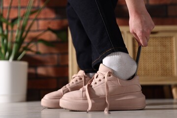  Woman putting on stylish sneakers with shoehorn indoors, closeup
