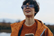 © SHOTPRIME STUDIO - Young woman lifestyle outdoors wearing glasses and orange jacket smiles happily while holding a smartphone. Background shows blurred natural landscape, concept of casual modern lifestyle and