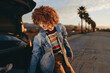© SHOTPRIME STUDIO - A woman with curly hair wearing a rainbow sweater and denim jacket smiles while reaching into the open trunk of a car on a sunny street lined with palm trees.