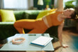 © Alliance - Health tracking tools including a scale and measuring tape sit on a table while a woman in orange fitness gear performs a side plank in the sunlit background.