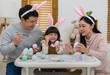 © geargodz - father, mother and daughter wearing bunny ears painting and decorating egg  together at home on easter day. happy family holiday activity concept