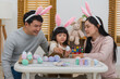 © geargodz - father, mother and daughter wearing bunny ears painting and decorating egg  together at home on easter day. happy family holiday activity concept