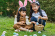 © geargodz - two happy child girl wearing bunny ears playing together and hunting for Easter eggs into basket on green grass in garden
