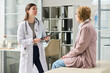© Mediaphotos - Young female doctor holding digital tablet speaking with senior woman sitting on examination table in medical office during consultation