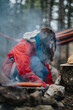 © qunica.com - A woman crouches by a smoky campfire cooking food in the woods. The scene shows casual outdoor camping with a hammock and camping gear nearby.