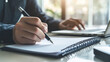 © Abderrahim - Close-up of professional man writing in notebook with pen at office desk with laptop, strategic planning and deep work concept
