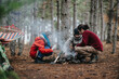 © qunica.com - Three friends sit around a small campfire in a pine forest on a mountain. The group tends the fire and prepares for a cozy outdoor evening together.