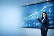 © JK_kyoto - Businesswoman pointing at rising graph on digital screen, confident professional woman in suit presenting financial growth and data analysis in modern office environment