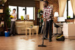 © DC Studio - Smiling african american man vacuums hardwood floor while Caucasian wife wipes shelves in modern living room. Diverse couple enjoys teamwork during household chores, maintaining cleanliness at home.
