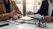 © Zahid - Two business professionals reviewing documents and using a calculator at a desk with a miniature house model, suggesting real estate, investment, or financial planning