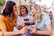 © Xavier Lorenzo - Multiracial female friends smiling with red wine at cafe table. Young women enjoying joyful social moment in European street. Lifestyle and social concept.