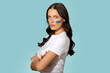 © Home-stock - Serious woman standing with her arms crossed and Ukrainian flag painted on her cheek, showing her support for Ukraine over blue background