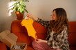 © wifesun - Pregnant woman sitting on a sofa unboxing a gift box, holding up a new yellow baby onesie and looking at it with a soft smile while preparing for the arrival of her child