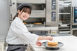 © buritora - A young Asian woman (pastry chef) in a chef's coat working in a confectionery workshop, cake shop kitchen, or bakery.