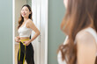 © KMPZZZ - Weight lose, loss concept. Slim body asian young woman hand use tape measuring around waistline in fit sports. Girl looking reflect in mirror at home. Healthy nutrition, fitness for wellbeing beauty.