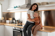 © sutulastock - Smiling mother in gray t-shirt sitting on kitchen counter and hugging her cheerful young son in brown outfit while spending happy family time together in modern bright kitchen