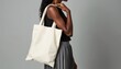 © Pete - Black woman holds simple canvas tote bag over shoulder. She wears black top and grey skirt. Minimalist accessory for shopping or daily use. Eco friendly bag. Neutral studio background.
