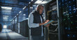 © Gorodenkoff - Data center technician inspects server racks, checks network cables, and uses a tablet to monitor hardware status. Rows of illuminated servers, digital infrastructure, and secure network operations.