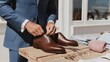 © DSimage - Man in blue suit tying laces on brown leather dress shoes displayed on wooden table with accessories and clothing.