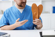 © New Africa - Smiling orthopedist in scrubs showing foot insoles at white desk in clinic, closeup