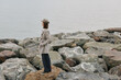 © SHOTPRIME STUDIO - A woman stands on rocky shore gazing at calm sea, wearing coat and hat. The overcast sky and muted colors create a peaceful, contemplative mood by the water landscape.