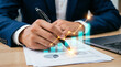 © maman - A businessman analyzing financial data on a document at his desk with a laptop nearby
