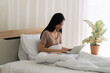 © kaew6566 - Young Asian woman working from home sitting on bed with laptop and notebook. Beautiful freelance female studying online or typing email in cozy bright bedroom. Remote work and education concept.