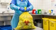 © QuiinL - A person in protective gear ties a yellow biohazard bag filled with medical waste in a laboratory setting.