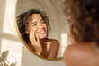 © EricMiguel - Curly - haired model smiling in sunlit bathroom, touching her freckled cheek while admiring her reflection in a round gold mirror wearing a white camisole