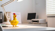 © Ann - Yellow rubber chicken on a white office desk under a lamp.